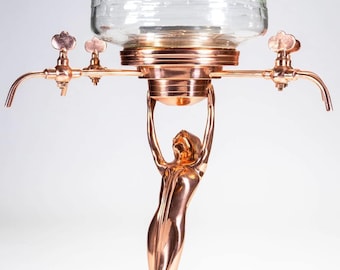 May include: A copper-toned absinthe fountain with a clear glass reservoir. The fountain features a decorative female figure supporting the base. It has two spouts with ornate handles, designed for serving absinthe.