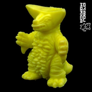 May include: A bright yellow dinosaur-like figurine with a textured surface. The figure has a large head crest, small arms, and a tail. The background is solid black. The text "STEKLU MORIKA" and a cat logo are in the upper right corner.