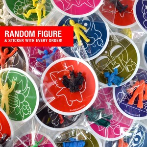 K&ouml;nnte beinhalten: Eine Sammlung bunter Aufkleber und kleiner Plastikfiguren. Die Aufkleber sind rund mit verschiedenen Cartoon-Designs und dem Text "@otakumerica". Der Text "RANDOM FIGURE & STICKER WITH EVERY ORDER!" wird auf einem roten Banner angezeigt.