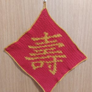 May include: A diamond-shaped, red crochet potholder with a gold border and a gold Chinese character design. It hangs from a loop at the top. The design is centrally placed on the potholder. The potholder is handmade.