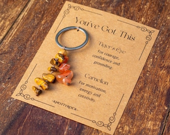 May include: A keychain featuring a silver ring and a collection of orange and yellow gemstones. The card reads "You've Got This" and details the properties of Tiger's Eye and Carnelian gemstones.