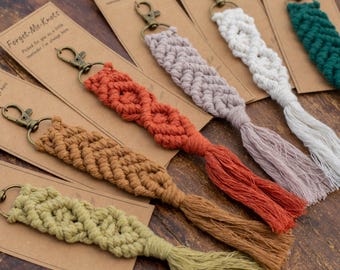 May include: A selection of macramé keyrings in shades of green, brown, orange, lavender, white, and teal. Each keyring features a metal clasp and a tassel. The keyrings are presented on small, rectangular cards with the text "Forget-Me-Knots."
