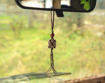 May include: A car rearview mirror hanging ornament featuring a rough, light-coloured stone encased in dark brown macrame cord. The design includes a single reddish-brown spherical bead above the stone and a thin, multi-strand brown cord tassel.