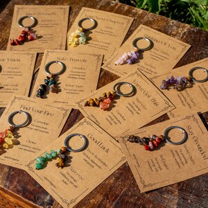 May include: A collection of keychains with various colorful gemstone beads attached to silver-tone rings. Each keychain is displayed on a small, tan card with text. The cards have decorative borders and are arranged on a wooden surface.