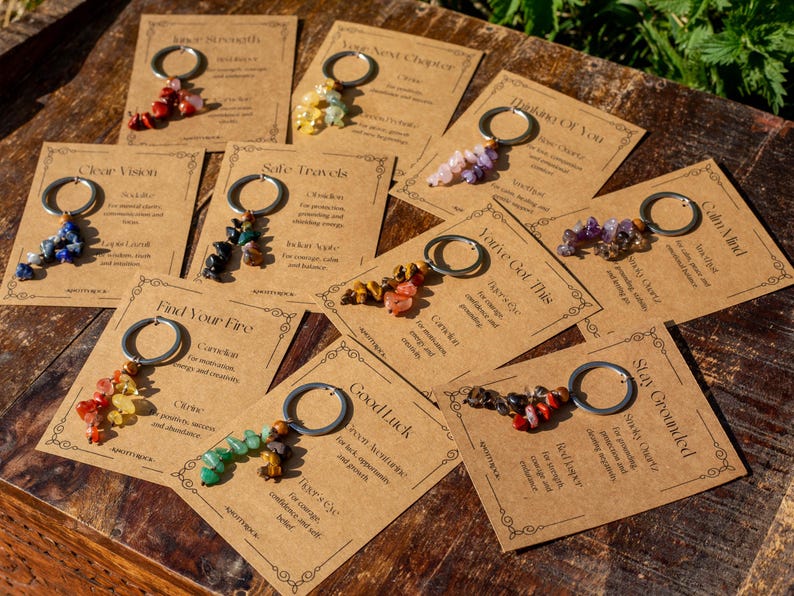 May include: A collection of keychains with various colorful gemstone beads attached to silver-tone rings. Each keychain is displayed on a small, tan card with text. The cards have decorative borders and are arranged on a wooden surface.