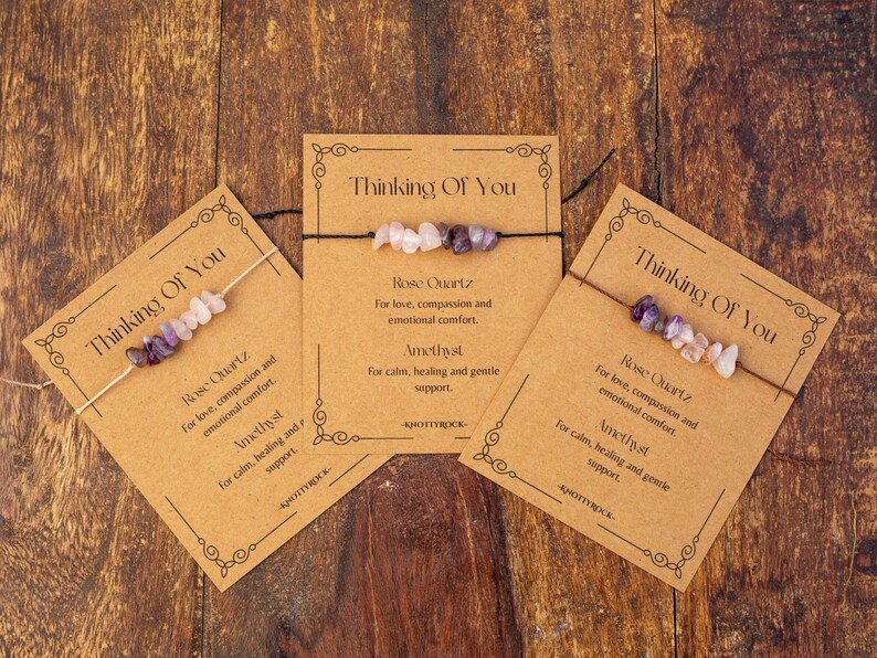 May include: Three gemstone bracelets on kraft paper cards with the text "Thinking Of You". Each card features a bracelet with rose quartz and amethyst beads. The cards are on a wooden surface.
