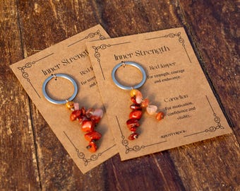 May include: Two keyrings with red jasper and carnelian beads, attached to silver rings, rest on brown cards. The cards read "Inner Strength" and include text about the stones' properties. The background is a wooden surface.