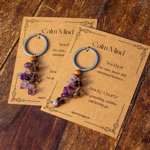 May include: Two keychains with amethyst and smoky quartz beads, attached to silver rings. The keychains are displayed on cards with the text "Calm Mind" and descriptions of the stones' properties. The background is a wooden surface.