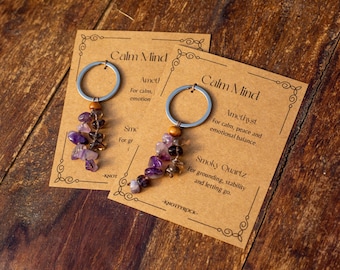 May include: Two keyrings featuring amethyst and smoky quartz beads, attached to silver rings. They are presented on cards with the text "Calm Mind" and descriptions of the stones' properties. The background is a wooden surface.