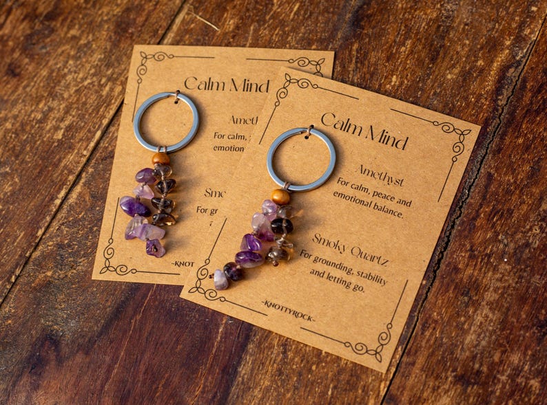 May include: Two keychains with amethyst and smoky quartz beads, attached to silver rings. The keychains are displayed on cards with the text "Calm Mind" and descriptions of the stones' properties. The background is a wooden surface.