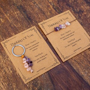 May include: Two cards with the text "Thinking Of You" on a wooden surface. One card has a keychain with pink and purple stones. The other card has a bracelet with similar stones. The cards also have text about Rose Quartz and Amethyst.