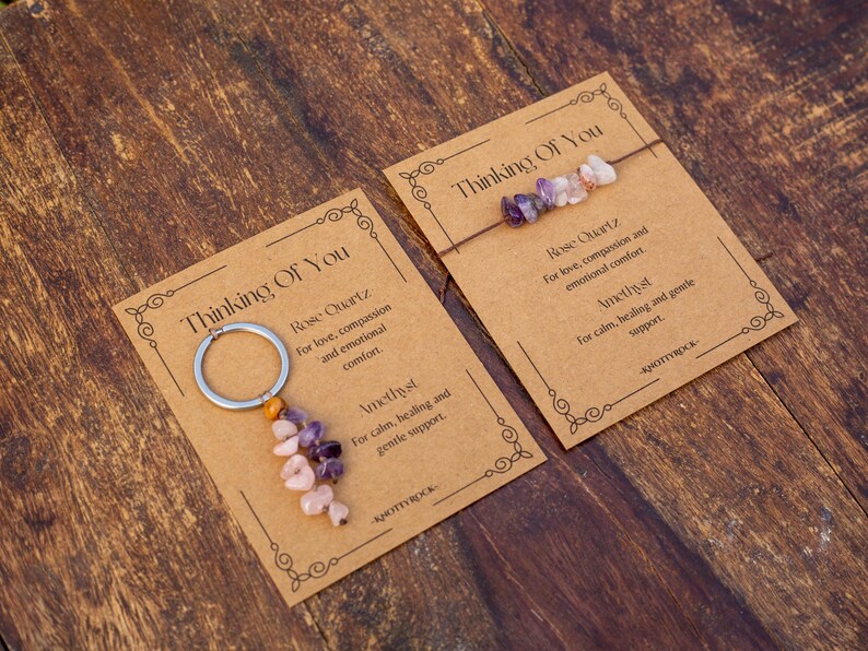 May include: Two cards with the text "Thinking Of You" on a wooden surface. One card has a keychain with pink and purple stones. The other card has a bracelet with similar stones. The cards also have text about Rose Quartz and Amethyst.