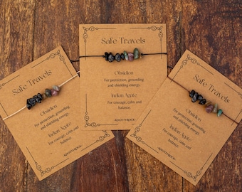May include: Three bracelets with obsidian and Indian agate beads are presented on brown cards. Each card reads "Safe Travels" and details the stones' properties. The cards are arranged on a wooden surface.