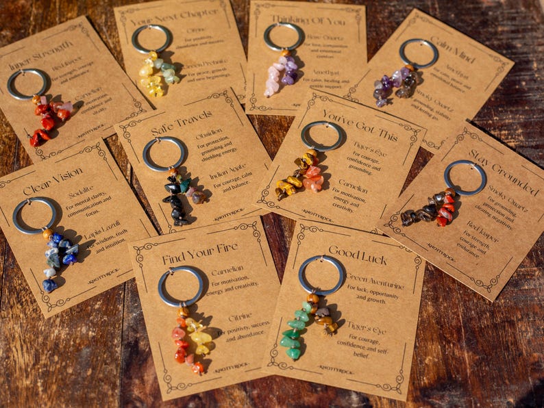 May include: A collection of keychains with various colorful gemstone clusters. Each keychain is attached to a small, brown card with text. The cards are arranged on a wooden surface, showcasing a variety of designs and purposes.