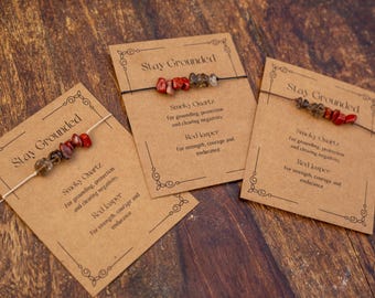 May include: Three gemstone bracelets on display, each presented on a brown card with the text "Stay Grounded." The bracelets feature a combination of smoky quartz and red jasper beads. The cards also list the properties of each stone.