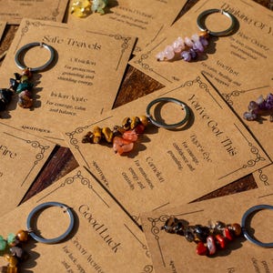 May include: Keychains with various gemstone beads attached to silver-colored rings are displayed on brown cards. The cards have text such as "Safe Travels" and "You've Got This." The keychains feature stones in colors like red, green, and purple.