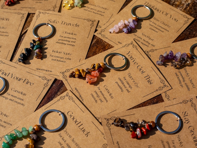 May include: Keychains with various gemstone beads attached to silver-colored rings are displayed on brown cards. The cards have text such as "Safe Travels" and "You've Got This." The keychains feature stones in colors like red, green, and purple.