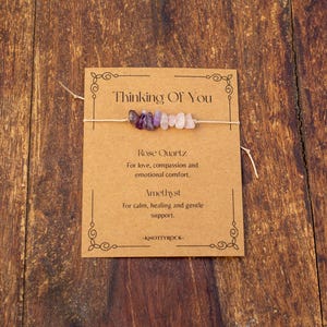 May include: A small bracelet with amethyst and rose quartz beads is displayed on a brown card. The card reads "Thinking Of You" and lists the properties of the stones. The background is a wooden surface.