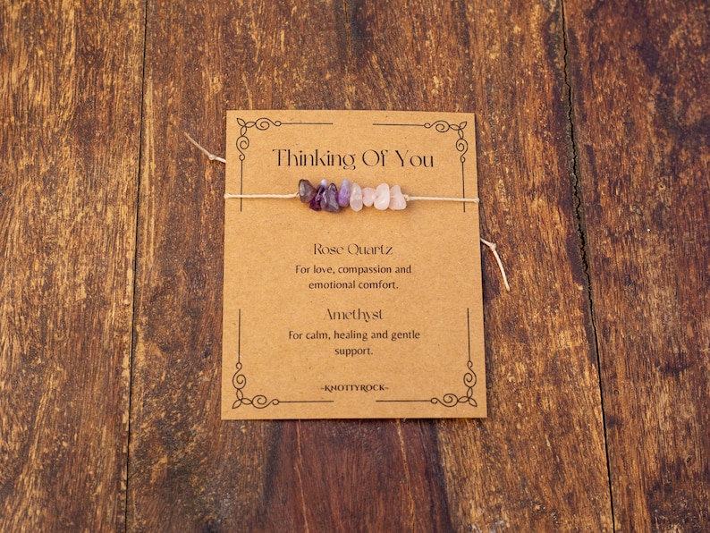 May include: A small bracelet with amethyst and rose quartz beads is displayed on a brown card. The card reads "Thinking Of You" and lists the properties of the stones. The background is a wooden surface.