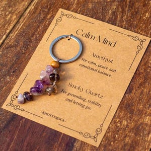 May include: A keychain with a silver ring and a cluster of purple and brown gemstones. The keychain is attached to a tan card with the words "Calm Mind" and descriptions of Amethyst and Smoky Quartz.