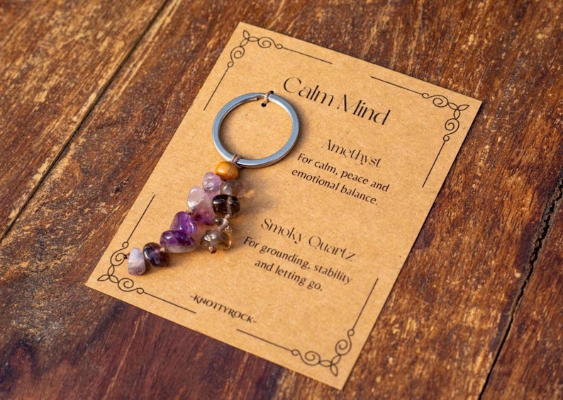 May include: A keychain with a silver ring and a cluster of purple and brown gemstones. The keychain is attached to a tan card with the words "Calm Mind" and descriptions of Amethyst and Smoky Quartz.