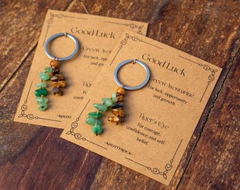 May include: Two keyrings with silver hoops and gemstone pendants. Each pendant features green and brown stones. The keyrings rest on cards with the words "Good Luck" and text about the stones' properties.