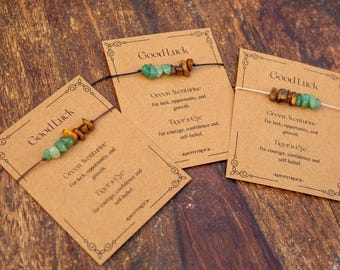 May include: Three bracelets on kraft paper cards labelled Good Luck. Each bracelet features small, irregular green aventurine and brown tiger's eye gemstone chips on a thin cord. The cards list the stone names and properties, ending with KNOTTYROCK.