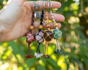 May include: Five keyrings with gemstones in various colours are held in a hand. Each gemstone is encased in a woven net, with wooden beads and a tassel of small stones. The gemstones include red, pink, brown, and green.
