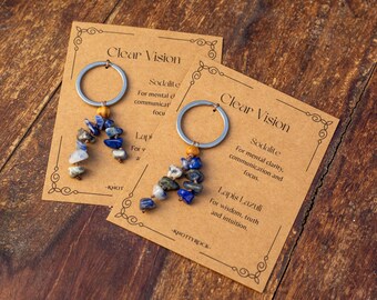 May include: Two keyrings with silver hoops and beaded gemstone details, presented on cards with the text "Clear Vision." The cards also list the properties of Sodalite and Lapis Lazuli. The background is a wooden surface.