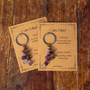 May include: Two keychains with amethyst and smoky quartz beads, attached to silver rings. Each keychain is displayed on a brown card with the text "Calm Mind" and descriptions of the stones' properties. The background is a wooden surface.