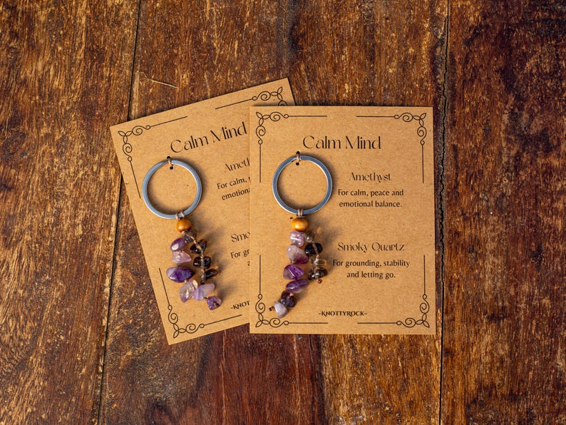 May include: Two keychains with amethyst and smoky quartz beads, attached to silver rings. Each keychain is displayed on a brown card with the text "Calm Mind" and descriptions of the stones' properties. The background is a wooden surface.