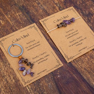 May include: Two cards with the text "Calm Mind" and product information. One card has a silver keychain with amethyst and smoky quartz stones. The other card has a string of amethyst and smoky quartz stones.