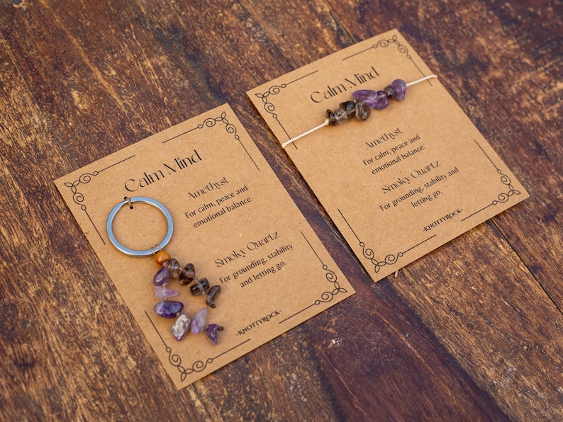 May include: Two cards with the text "Calm Mind" and product information. One card has a silver keychain with amethyst and smoky quartz stones. The other card has a string of amethyst and smoky quartz stones.