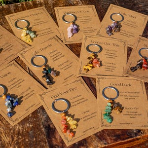 May include: A collection of keychains with various gemstone clusters attached to silver rings, each displayed on a brown card with text. The cards have phrases like "Clear Vision" and "Good Luck". The keychains are on a wooden surface.