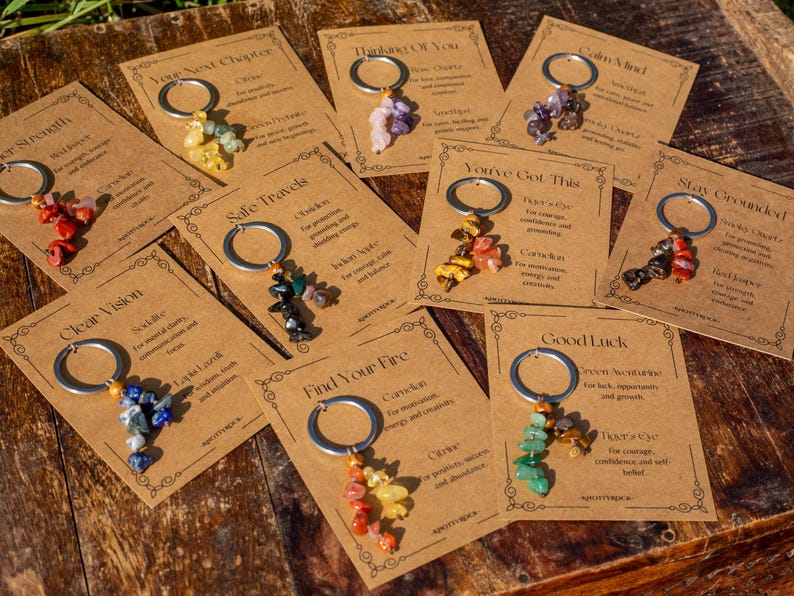 May include: A collection of keychains with various gemstone clusters attached to silver rings, each displayed on a brown card with text. The cards have phrases like "Clear Vision" and "Good Luck". The keychains are on a wooden surface.