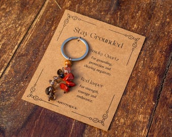 May include: A keychain with a silver ring and a cluster of beads in shades of brown, red, and clear. The keychain is displayed on a brown card with the text "Stay Grounded" and information about Smoky Quartz and Red Jasper.
