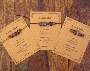 May include: Three bracelets with amethyst and smoky quartz beads are displayed on brown cards. The cards read "Calm Mind" and list the stones' properties. The background is a wooden surface.