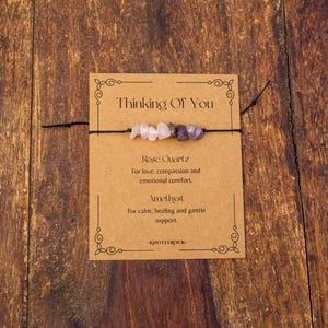 May include: A handmade bracelet featuring rose quartz and amethyst beads strung on a black cord, presented on a brown card. The card reads "Thinking Of You" and describes the stones' properties. The background is a wooden surface.