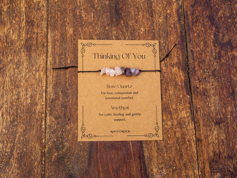 May include: A handmade bracelet featuring rose quartz and amethyst beads strung on a black cord, presented on a brown card. The card reads "Thinking Of You" and describes the stones' properties. The background is a wooden surface.