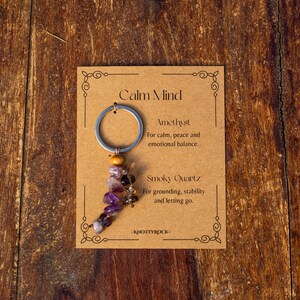 May include: A keychain with a silver ring and a strand of amethyst and smoky quartz beads. The card reads "Calm Mind" and lists the properties of the stones. The background is a wooden surface.