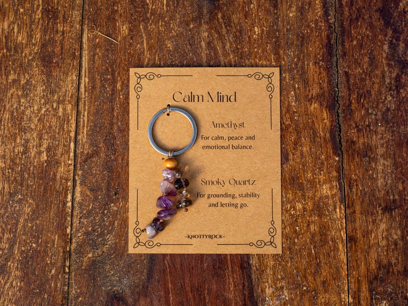 May include: A keychain with a silver ring and a strand of amethyst and smoky quartz beads. The card reads "Calm Mind" and lists the properties of the stones. The background is a wooden surface.