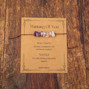 May include: A bracelet with a string of small, colorful gemstones, including purple amethyst and pink rose quartz, is displayed on a brown card. The card reads "Thinking Of You" and lists the properties of the stones.