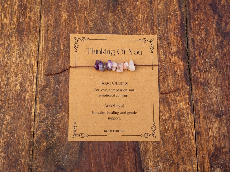 May include: A bracelet with a string of small, colorful gemstones, including purple amethyst and pink rose quartz, is displayed on a brown card. The card reads "Thinking Of You" and lists the properties of the stones.