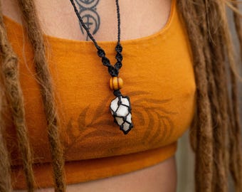 May include: A black macrame necklace with a white crystal pendant and a wooden bead. The necklace is displayed against an orange top with a fern design. The person has long, brown dreadlocks.