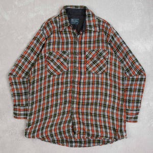 Pode incluir: Uma camisa de manga comprida com botões e um padrão xadrez em vermelho, preto, branco e verde. A camisa tem dois bolsos no peito e uma gola preta. A etiqueta diz "The Men's Shop".
