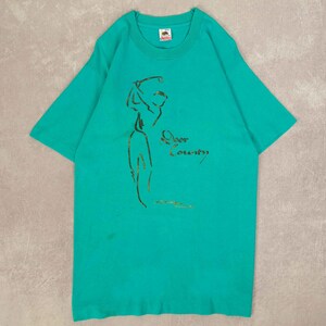 以下が含まれることがあります： ターコイズ色のTシャツ。金色の輪郭でゴルフをする人のイラストが描かれています。「Door County」の文字が筆記体でプリントされています。柔らかい素材でできており、半袖です。