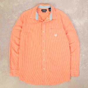 May include: A long-sleeved shirt in orange and white check pattern, featuring a button-down design and a chest pocket. The collar has a light blue lining, and the brand name "CHAPS" is visible.