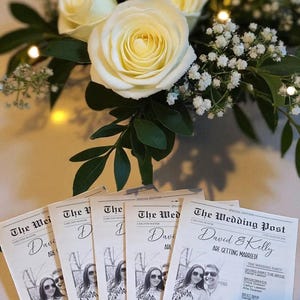 May include: A wedding reception scene featuring a bouquet of white roses and small white flowers. Several copies of a wedding newspaper, "The Wedding Post," are displayed, with photos and event details. The newspaper's headline reads "David & Kelly Are Getting Married!"