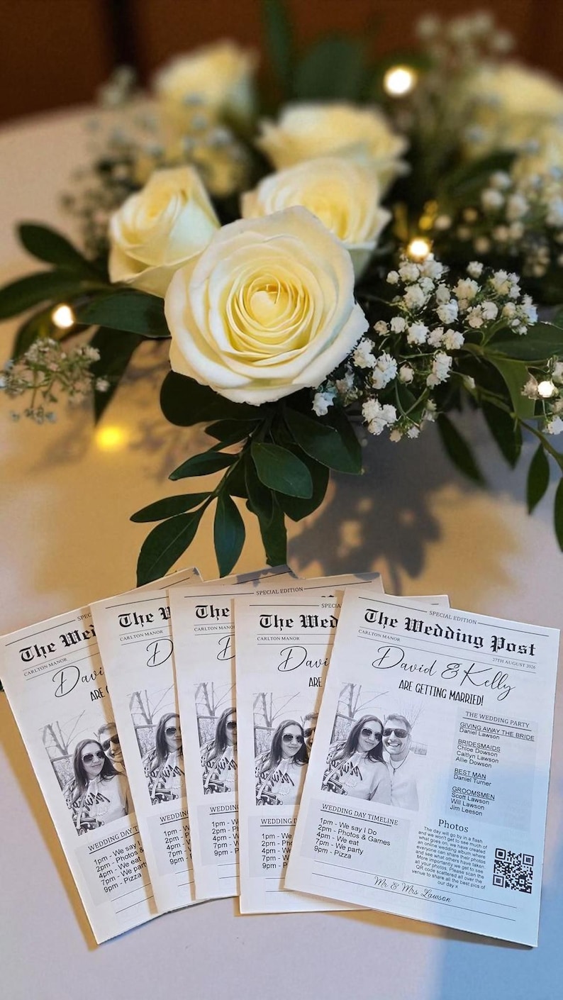 May include: A wedding reception scene featuring a bouquet of white roses and small white flowers. Several copies of a wedding newspaper, "The Wedding Post," are displayed, with photos and event details. The newspaper's headline reads "David & Kelly Are Getting Married!"