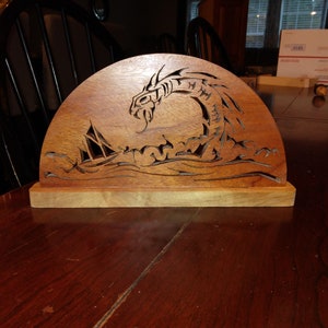 May include: A wooden dragon sculpture with intricate details, carved in a half-circle shape. The dragon is depicted with its head raised and its wings spread out, as if it is about to take flight. The sculpture is made of a light brown wood and has a natural finish.